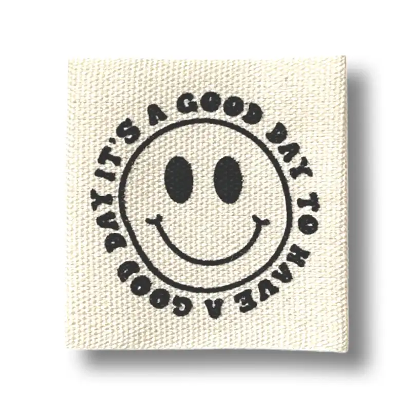 quadratisches Baumwoll-Label mit schwarzem Smiley in der Mitte. Runder Schriftzug um das Smiley "it`s a good day to have a good day"